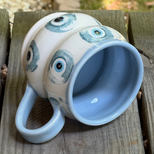 Load image into Gallery viewer, Ceramic mug with blue interior and decorative eyes on exterior, placed on wooden surface.