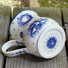 Load image into Gallery viewer, White mug with blue and green designs on a wooden surface outdoors
