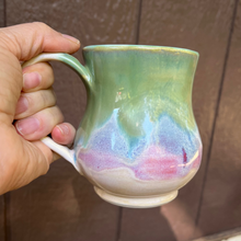 Load image into Gallery viewer, Hand holding a ceramic mug with a multicolored design against a brown wall.
