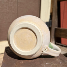 Load image into Gallery viewer, Beige ceramic mug with a green and beige handle on a metal surface
