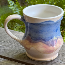 Load image into Gallery viewer, Ceramic mug with blue and pink glaze on a wooden surface with greenery in the background
