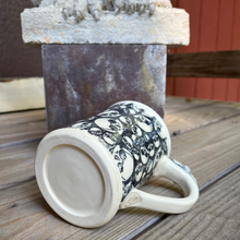 Load image into Gallery viewer, White ceramic mug with black patterns on a wooden surface, with a stone sculpture in the background.
