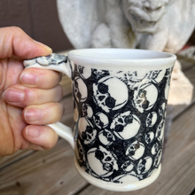 Load image into Gallery viewer, Person holding a mug with skull pattern in front of a gargoyle statue.
